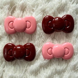 Puede incluir: Cuatro adornos en forma de lazo, en rosa claro y burdeos oscuro, sobre una superficie blanca texturizada. Los lazos brillantes tienen un diseño clásico, adecuados para manualidades o accesorios. Miden aproximadamente 2 cm de largo.