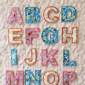 May include: A complete alphabet of fabric letters, each featuring a floral design in diverse colours. The letters are outlined in orange and blue, and are arranged on a white, textured surface. The letters are in uppercase.