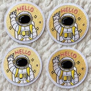 Peut inclure: Quatre écussons brodés circulaires représentant un astronaute en combinaison blanche et jaune faisant un signe de la main. Le fond est jaune avec de petites étoiles et le mot HELLO brodé en lettres roses au-dessus du casque.