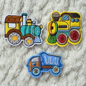 May include: Three embroidered patches of transport vehicles: a train with brown, teal, and yellow details, a yellow train with red wheels, and a blue dump truck with orange accents. The patches are on a white, textured surface.