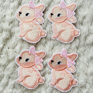 Op de afbeelding: Vier geborduurde patches met een zittend konijn met een roze strik. De konijnen zijn licht perzik met donkerdere perzik accenten en een witte omtrek. Op een wit, getextureerd oppervlak.