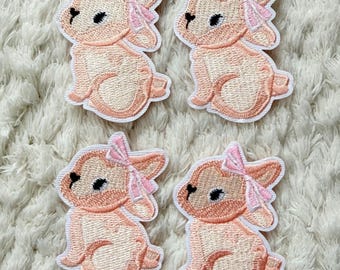 May include: Four embroidered patches featuring a sitting bunny with a pink bow. The bunnies are light peach with darker peach accents and a white outline. The patches are on a white, textured surface.