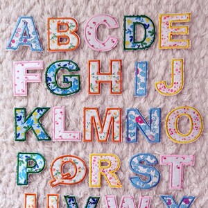 以下が含まれることがあります： さまざまな色の花柄が施された、刺繍されたアルファベットの文字のコレクション。文字はさまざまな色で縁取られ、柔らかく質感のある薄いピンク色の表面に配置されています。