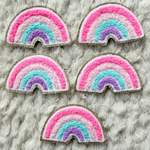Könnte beinhalten: Fünf bunte Regenbogen-Aufnäher mit rosa, blauen und lila Streifen und einem goldglitzernden Rand. Die Aufnäher sind auf einer weißen, strukturierten Oberfläche angeordnet. Diese Aufnäher sind ideal, um Kleidung oder Accessoires einen Hauch von Laune zu verleihen.