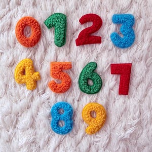 Può includere: Una collezione di numeri colorati e strutturati da 0 a 9 disposti su una superficie bianca e soffice. Ogni numero è di un colore diverso, tra cui arancione, verde, rosso, blu, giallo e altro ancora. I numeri sembrano essere fatti di un materiale morbido.