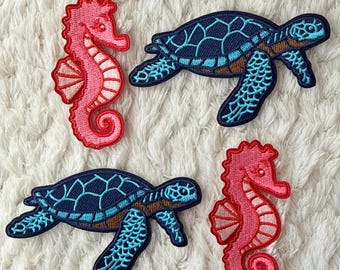 May include: Four embroidered patches featuring sea creatures. Two pink seahorses with white and red details, and two blue sea turtles with brown and turquoise accents. The patches are on a white, textured background.