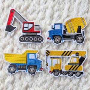 Könnte beinhalten: Vier gestickte Aufnäher mit Baumaschinen: ein roter Bagger, ein blauer Betonmischwagen, ein gelber Muldenkipper und ein gelber Kranwagen. Jeder Aufnäher hat einen weißen Rand auf einem weißen, strukturierten Hintergrund.