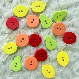 Può includere: Una collezione di piccoli bottoni a due fori a forma di frutta, tra cui limoni gialli, cerchi arancioni, mele rosse e pere verde chiaro. Questi bottoni piatti e colorati sono sparsi, mostrando diverse sagome di frutta.