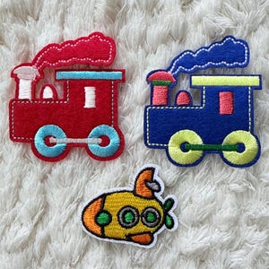 Può includere: Tre toppe di stoffa colorate: un treno rosso, un treno blu e un sottomarino giallo. I treni hanno dettagli bianchi e fumo blu o rosso. Il sottomarino ha eliche e oblò verdi.