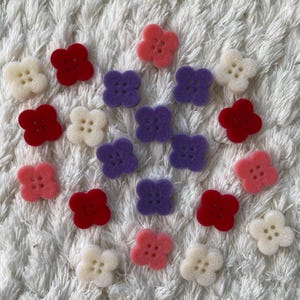 Può includere: Una collezione di bottoni a forma di fiore in vari colori, tra cui rosso, rosa, viola e crema. I bottoni sono disposti su una superficie bianca e strutturata, creando un motivo visivamente accattivante. Ogni bottone ha quattro petali e piccoli fori per cucire.