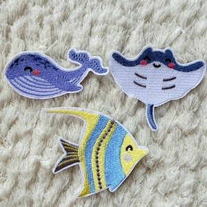 Peut inclure: Trois écussons brodés représentant des créatures marines : une baleine bleue, une raie manta blanche et bleue, et un poisson jaune et bleu. Chaque écusson a un visage souriant, sur fond blanc texturé.