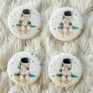 May include: Four round white buttons, each featuring a printed illustration of an astronaut sitting on a small planet. The design includes small stars and orange dots scattered across the surface, with two holes centred on the astronaut's torso.