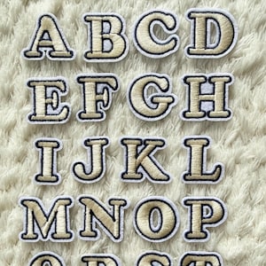 Könnte beinhalten: Gestickte Alphabet-Patches mit den Buchstaben A bis Z. Jeder Buchstabe ist in einer Serifenschrift mit goldfarbener Füllung und schwarzem Rand gehalten. Die Patches sind auf einer weißen, strukturierten Oberfläche angeordnet.