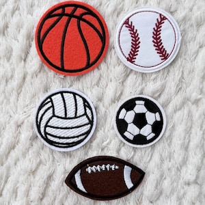 Può includere: Una collezione di toppe a tema sportivo. Include una toppa da basket rossa, una toppa da baseball bianca con cuciture rosse, una toppa da pallavolo bianca con dettagli neri, una toppa da calcio bianca e nera e una toppa da football americano marrone.