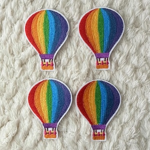 Pode incluir: Quatro patches bordados em forma de balões de ar quente. Cada balão apresenta um design de arco-íris com listras vermelhas, laranja, amarelas, verdes, azuis e roxas. Os balões têm uma borda branca e uma cesta roxa.