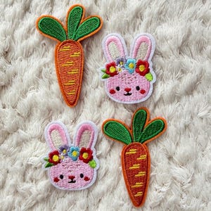 Peut inclure: Quatre écussons brodés : deux carottes et deux visages de lapin roses. Les carottes sont oranges avec des fanes vertes. Les visages de lapin ont des oreilles roses et portent des couronnes de fleurs. Sur fond blanc texturé.