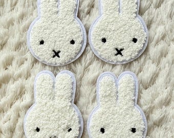 May include: Four white bunny patches with fluffy texture, black eyes, and a black 'X' for a mouth. Each bunny is outlined with a light blue border. The patches are arranged on a white, textured surface.