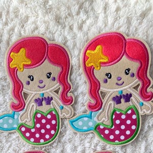 Könnte beinhalten: Vier Filz-Meerjungfrauen-Patches mit rosa Haaren, einem gelben Stern, einem lila Oberteil und einem rosa-weiß gepunkteten Schwanz.