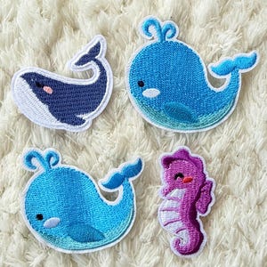 May include: Four embroidered patches featuring sea creatures. There are three blue whales and one purple seahorse. The whales have white bellies and dark blue bodies. The seahorse is purple with white accents and a smiling face.