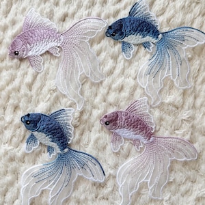 May include: Embroidered goldfish patches in blue and purple hues, with sheer, delicate tail fins. The patches display detailed stitching, highlighting the fish's scales and fins. They are set against a textured, white background.