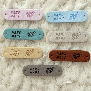 Könnte beinhalten: Sieben rechteckige Etiketten in verschiedenen Pastellfarben, jeweils mit den Worten "HAND MADE" und einem Garnknäuel- und Nadeldesign. Die Etiketten sind in Rosa, Blau, Grün, Beige, Gelb, Braun und Grau gehalten.