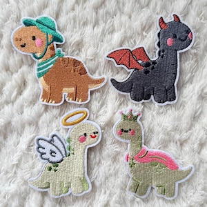 May include: Four embroidered dinosaur patches. One is brown with a green hat and scarf, another is black with red wings, a third is light green with a halo and wings, and the last is light green with a pink shawl and crown.