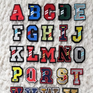 May include: A collection of embroidered alphabet patches in a variety of colours, such as red, blue, green, and yellow. Each letter has a contrasting outline and a sporty, collegiate design. The patches are displayed on a white, textured surface.
