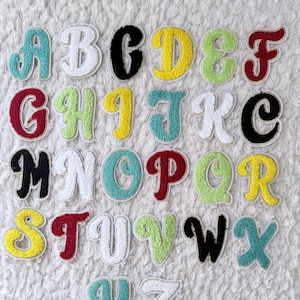 May include: A collection of colorful, embroidered alphabet letters. Each letter is a different color, including teal, white, black, yellow, green, and red. The letters are on a white, textured background.