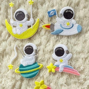 Może przedstawiać: Haftowane naszywki przedstawiające kreskówkowych astronautów w różnych scenach kosmicznych. Jeden astronauta huśta się na księżycu, inny jedzie na rakiecie, a inni są na planecie, tęczy i trzymają balony. Każdy astronauta ma biały skafander kosmiczny.