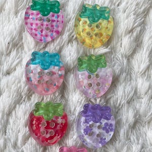 Könnte beinhalten: Sechs erdbeerförmige Knöpfe mit grünen Stielen. Die Knöpfe zeigen verschiedene Designs, darunter rosa Karomuster, gelb mit orangefarbenen Punkten und lila mit Blumenmustern. Jeder Knopf hat drei oder vier Löcher zum Annähen.