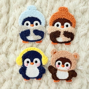 May include: Four embroidered penguin patches in different colours. Each penguin has a hat or hood, including blue, orange, and yellow. The penguins have white bellies, black eyes, and orange feet. The patches are on a white, textured surface.