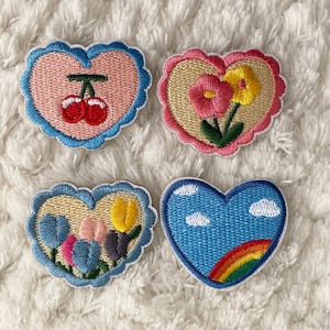 Può includere: Quattro toppe ricamate da stirare a forma di cuore. Ogni toppa presenta un design diverso: ciliegie, fiori, tulipani e un arcobaleno con nuvole. Le toppe sono tutte su una superficie bianca e soffice.