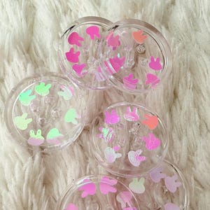 Könnte beinhalten: Klare, runde Untersetzer mit Hasen-Konfetti in Rosa-, Grün- und Orangetönen. Die Untersetzer bestehen aus transparentem Kunststoff und liegen auf einer flauschigen, weißen Oberfläche.