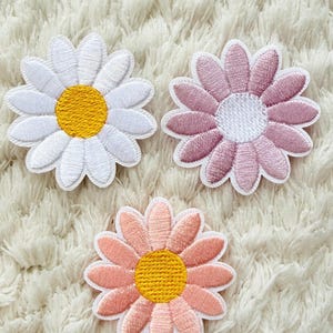 Può includere: Tre toppe ricamate a forma di margherita in bianco, rosa e pesca con centri gialli. Le toppe sono su uno sfondo bianco strutturato, che mostra le cuciture dettagliate e i colori vivaci dei disegni floreali.