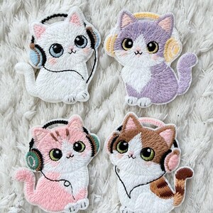 Könnte beinhalten: Vier gestickte Katzenaufnäher, jeweils mit Kopfhörern. Die Katzen sind weiß, lila, rosa und braun, mit detaillierten Gesichtern und farbigen Kopfhörern. Auf einer weißen, strukturierten Oberfläche.