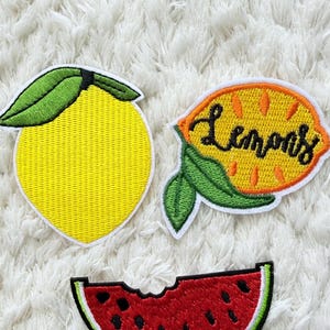 Könnte beinhalten: Gestickte Aufnäher mit Fruchtdesigns. Enthält eine gelbe Zitrone, eine Zitrone mit dem Wort "Lemons" in Schrift und eine Wassermelonen-Scheibe. Die Aufnäher haben einen weißen Rand und befinden sich auf einem weißen, strukturierten Hintergrund.