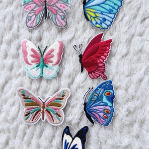 May include: A collection of embroidered butterfly patches in various colors, including pink, blue, red, and brown. Each patch features detailed stitching and unique wing patterns. The patches are arranged on a white, textured surface.