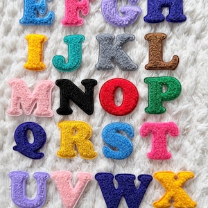 Puede incluir: Una colección de letras del alfabeto bordadas y coloridas. Cada letra es de un color diferente, incluyendo rojo, verde, azul, amarillo, rosa y morado. Las letras están dispuestas sobre una superficie blanca y texturizada, creando una exhibición vibrante y juguetona.