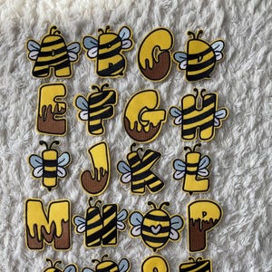 Può includere: Un set di 26 toppe da stirare con l'alfabeto in un design a forma di ape. Ogni lettera è gialla con strisce nere e un viso di ape. Alcune lettere hanno miele marrone che cola.