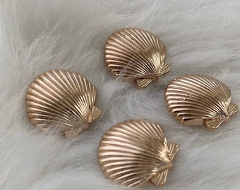 May include: Four gold-colored seashell-shaped hair clips. The clips are arranged on a white fluffy surface.
