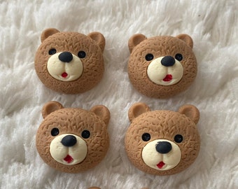 May include: Five brown bear face buttons with black eyes and a red mouth. The buttons are on a white fluffy surface.