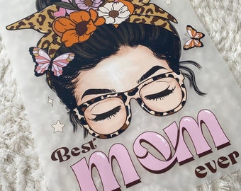May include: A white background with a pink and brown graphic of a woman with a messy bun wearing leopard print glasses and a headband with flowers and leopard print. The text "Best mom ever" is written in pink and brown letters.