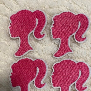 Peut inclure: Quatre patchs thermocollants en tissu rose en forme de silhouette de femme avec une queue de cheval. Les patchs ont un contour blanc.