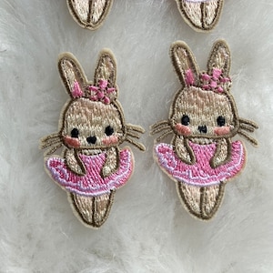 Könnte beinhalten: Vier braune gestickte Hasen-Patches mit rosa Tüllröcken und Schleifen. Die Hasen haben schwarze Augen und rosa Wangen.