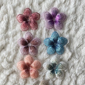 Può includere: Sette toppe di fiori ricamati in vari colori, tra cui rosa, viola, blu e bianco. I fiori hanno un design a cinque petali con un piccolo centro.