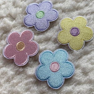 Può includere: Quattro toppe di fiori ricamate in diversi colori. Le toppe sono viola, rosa, giallo e blu. Ogni toppa ha un cerchio più piccolo al centro.