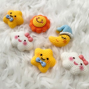 May include: Six colourful, small, plush toys with happy faces. The toys are shaped like a star, a sun, a moon, and two clouds. The toys are on a white fluffy surface.