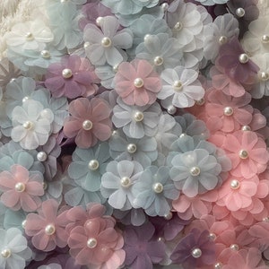 Könnte beinhalten: Eine Sammlung von pastellfarbenen Stoffblumen mit Perlenzentren. Die Blumen sind in einem zufälligen Muster angeordnet und haben alle unterschiedliche Größen.