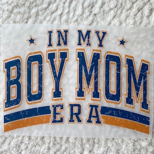 May include: A white iron-on transfer with blue and orange text that reads "In My Boy Mom Era" with a star on either side of the text.