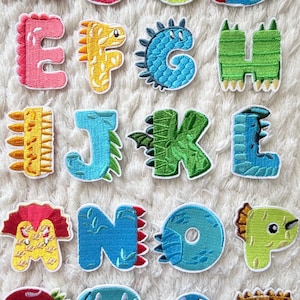 Könnte beinhalten: Gestickte Buchstaben-Patches im Dinosaurier-Design in verschiedenen Farben. Jedes Buchstaben-Patch ist als ein anderer Dinosaurier gestaltet. Ideal zum Aufnähen auf Kleidung oder Accessoires.